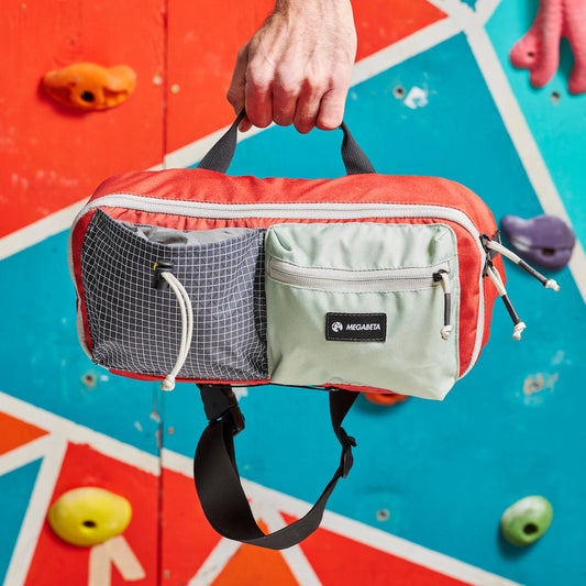 A climbing sling bag with red, gray, and black colors is displayed in front of a climbing wall with handholds. The bag features multiple compartments and a strap.